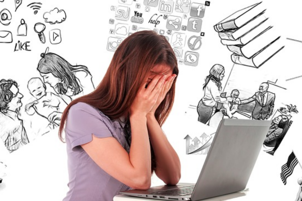 Young female seated facing open laptop with face in hands expressing overwhelm thinking about multitasking